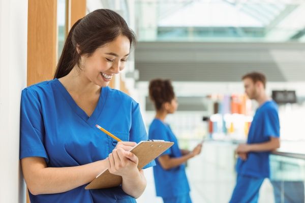 Female nurse smiling and making notes on a clipboard in a hospital setting with two other nurses conversing behind her