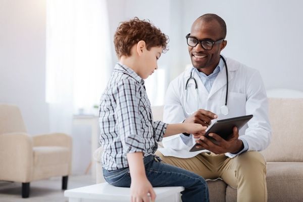 Male family nurse practitioner using a tablet to perform health assessment on a boy while sitting on a couch