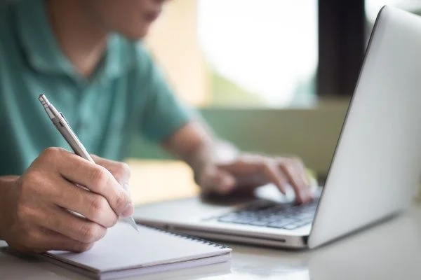 Close-up of male online nursing student studying on a laptop and taking notes in a notebook