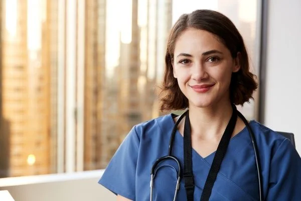 Young female nurse smiling in blue scrubs with a city view outside the window behind her