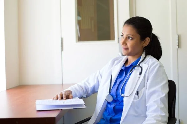 Female nurse practitioner sitting in her office and resting her hand on an open notebook while looking upwards and smiling softly