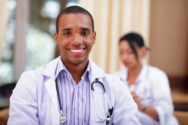 Smiling male nurse practitioner in a white lab coat with female nurse practitioner in the background