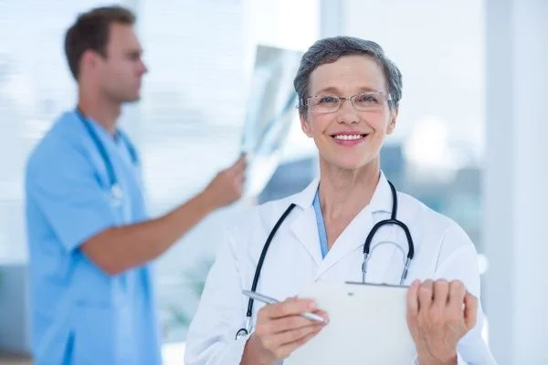 Smiling mature female nurse practitioner in a white lab coat with a male nurse examining an X-ray in the background