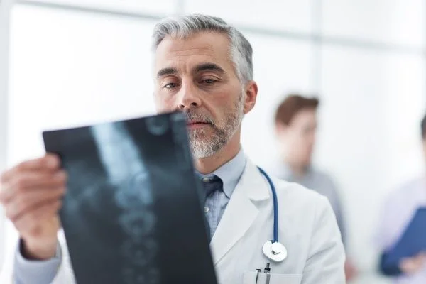 An older male doctor with grey hair and a cropped beard closely examining an X-ray