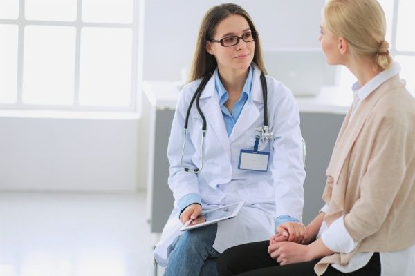 Young female nurse practitioner in a white lab coat listening patiently to her female patient explaining her health concerns while grasping her hand