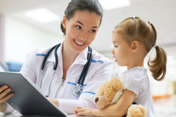 Smiling female nurse practitioner holding a clipboard and chatting to a young girl holding a teddy bear