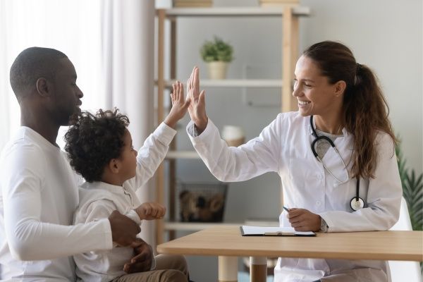 Female nurse practitioner high-fiving a young boy being held by his father in her office