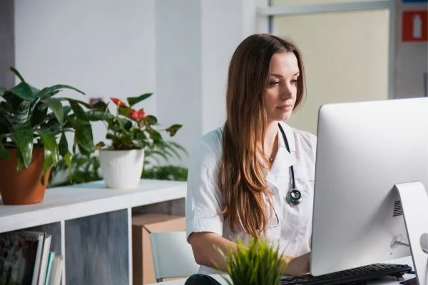 Female nurse practitioner wearing a white lab coat and stethoscope doing work at an office computer