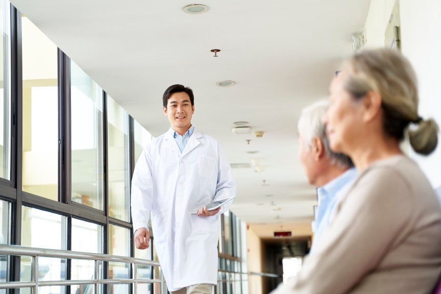 Male nurse practitioner walking down a hospital corridor in front of two elderly patients