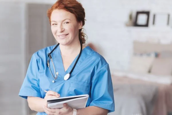 Smiling middle-aged female nurse in blue scrubs holding a notebook and pen